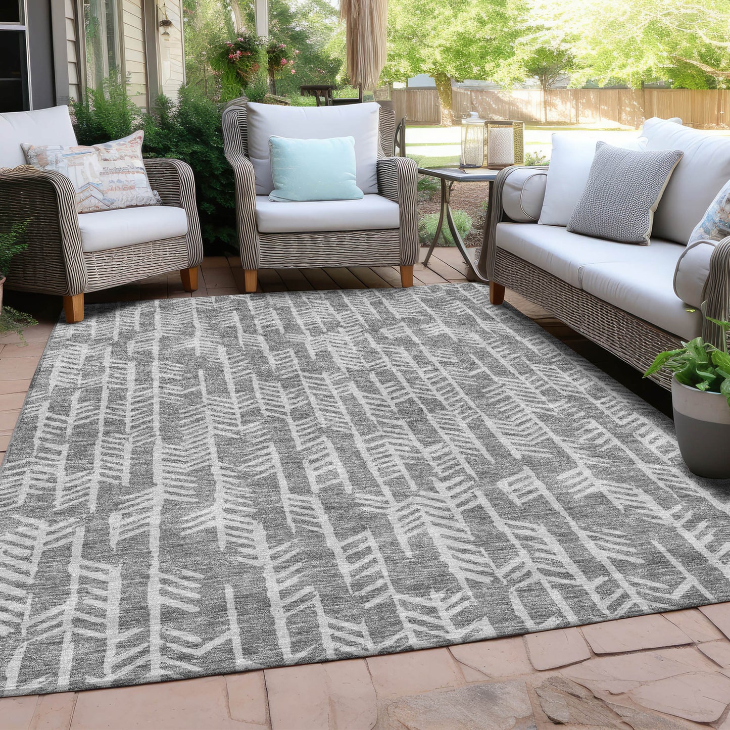 An all-weather gray and white outdoor rug with a hand-carved geometric pattern, placed on a patio under a covered porch. The rug is positioned under outdoor wicker furniture.